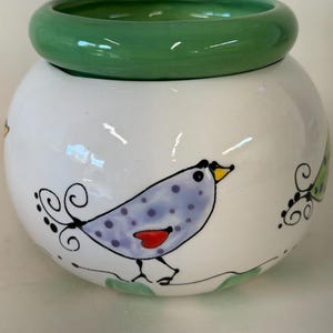 May include: A round white ceramic bowl featuring a hand-painted light purple bird with dark purple polka dots, a yellow beak, and a red heart-shaped wing. The bowl has a thick, smooth, solid green rim and a green interior finish.