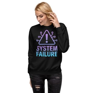 Puede incluir: Sudadera negra con un gráfico que dice "SYSTEM FAILURE". El diseño presenta un símbolo de advertencia morado y azul con un signo de exclamación en el interior. La sudadera es de estilo cuello redondo.