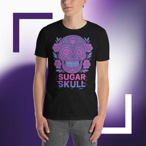Puede incluir: Camiseta negra con un diseño de calavera de azúcar vibrante. La calavera está adornada con patrones florales rosas y azules, un corazón y el texto "SUGAR SKULE" en degradado. El diseño está centrado en la camiseta.