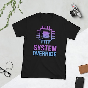Può includere: T-shirt nero con grafica di chip per computer viola e blu e la scritta "SYSTEM OVERRIDE". Il testo e la grafica sono centrati. La maglietta è a maniche corte.