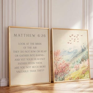 Puede incluir: Dos impresiones artísticas enmarcadas. Una presenta el texto de Mateo 6:26 en un marco dorado. La otra impresión representa un paisaje en acuarela con montañas, árboles y pájaros en un marco dorado. La obra de arte tiene una paleta de colores pastel.