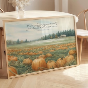 May include: A framed print featuring a pumpkin patch landscape with a field of orange pumpkins. The artwork includes a quote: "Rejoice always, pray continually, give thanks in all circumstances." The print is in a light wooden frame.
