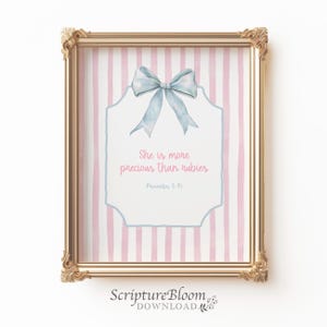 Peut inclure: Impression encadrée dans un cadre doré orné. L'impression présente un nœud bleu clair et le texte "She is more precious than rubies" sur un fond blanc avec des rayures verticales roses et blanches. Le texte "Proverbes 3:15" est également inclus.