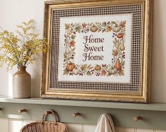 May include: A framed cross-stitch sampler featuring the text Home Sweet Home surrounded by a decorative border of pumpkins, acorns, and autumn leaves. The artwork is set within a gold-toned frame with a brown and white gingham fabric matting.