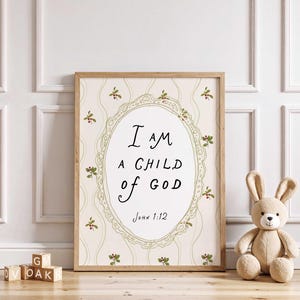 May include: A framed print with the text "I AM A CHILD OF GOD" in black script. The print has a cream-colored background with a floral design and is framed in light brown wood. A stuffed bunny and wooden blocks are in the foreground.