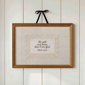 May include: A rectangular gold frame containing a beige fabric background with thin vertical stripes. Centered is a white lace-trimmed fabric piece featuring the black text: Be still, and know that I am God. Psalm 46:10. A black ribbon hangs from the top.