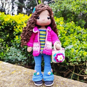 Peut inclure: Une poupée en crochet aux cheveux bouclés bruns, portant une veste rose, un chemisier rayé, un jean bleu avec des accents de fleurs et tenant un sac en crochet blanc avec un motif floral.