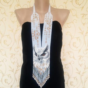 May include: A beaded owl necklace with a light blue and white background. The owl has detailed feathering and brown eyes. The necklace has a fringe of beads at the bottom and geometric patterns at the top.