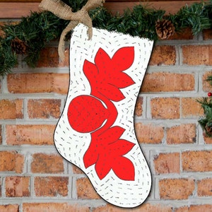 May include: A white Christmas stocking with red holly leaf and berry designs. The stocking is hanging on a brick fireplace mantel with a green garland.