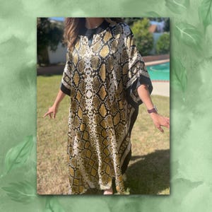 May include: A long, flowing caftan in a snakeskin pattern of tan, black, and cream. The caftan has a V-neck and wide sleeves with black trim. The model is standing on grass in front of a swimming pool.