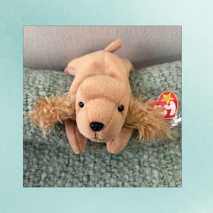May include: A tan plush dog toy with floppy ears and a black nose. The toy has a red heart-shaped tag with a star and text. The dog is lying on a green textured surface.