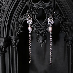 May include: A pair of silver-toned earrings with a knot design at the top, featuring a dangling chain and a pinkish-white pearl. The earrings are displayed against a dark, gothic-style background.