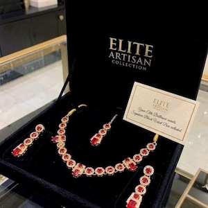 May include: A gold-toned jewelry set with red gemstones, including a necklace, earrings, and a headpiece, presented in a black velvet box. The box is branded with "ELITE ARTISAN COLLECTION". A card reads "Your Elite Brilliance awaits. Sapphire Black Velvet Box included."