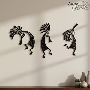 May include: Three black metal wall art pieces depicting stylized figures with headdresses and musical instruments. The figures are in various poses, with intricate patterns and details. The art is mounted on a light-coloured wall.