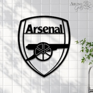 May include: Black metal wall art featuring the Arsenal logo. The logo includes the word "Arsenal" in a bold font above a cannon design, all within a shield shape. The art is mounted on a white tiled wall.