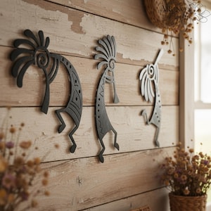 May include: Three black metal Kokopelli wall art pieces are mounted on a weathered, light-coloured wooden wall. The Kokopelli figures have stylised bodies and headdresses. The image includes the text "Aruno Gifts" in the upper right corner.