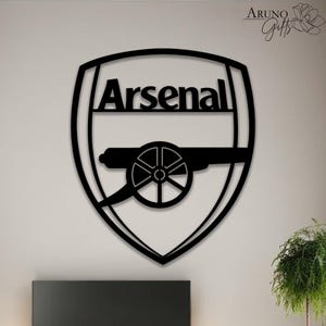 May include: Black metal Arsenal logo wall art. The design features the word "Arsenal" above a cannon within a shield shape. The artwork is suitable for home decor and fan merchandise.