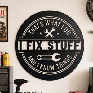 May include: A circular black metal wall sign featuring the text THAT'S WHAT I DO, I FIX STUFF, AND I KNOW THINGS. The design includes a pair of crossed wrenches at the top and a single wrench icon in the center, all cut from a solid black panel.