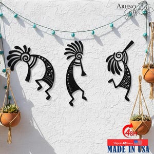 May include: Three black metal Kokopelli wall art pieces are mounted on a textured white wall. The Kokopelli figures are in various dancing poses. Hanging planters with succulents and a string of turquoise lights decorate the wall. The text "MADE IN USA" is visible.