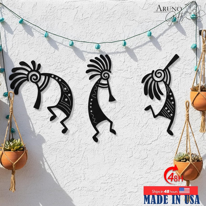 May include: Three black metal Kokopelli wall art pieces are mounted on a textured white wall. The Kokopelli figures are in various dancing poses. Hanging planters with succulents and a string of turquoise lights decorate the wall. The text "MADE IN USA" is visible.