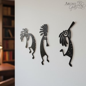 May include: Three black metal Kokopelli wall art pieces are mounted on a white wall. Each figure has a distinct design, with details like headdresses and flute-like instruments. The figures are in various poses, adding a dynamic feel to the decor.