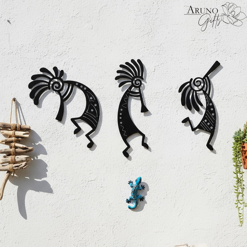 May include: Three black metal Kokopelli wall art pieces are mounted on a white wall. The Kokopelli figures have stylized features and are in different poses. A small blue lizard and driftwood decor are also on the wall. The brand name "Aruno Gifts" is in the top right corner.