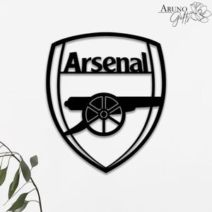 May include: Black metal Arsenal logo. The logo is a shield shape with the word "Arsenal" at the top. Below the word is a cannon. The logo is a popular symbol for fans of the Arsenal football club.