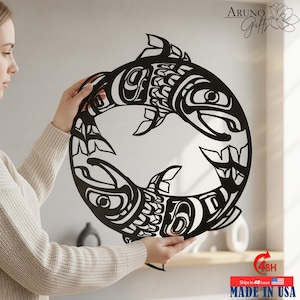 May include: A black metal wall art piece featuring a circular design of two stylized fish, with intricate tribal patterns. The artwork is held by a person. The text "Aruno Gifts" is in the upper right corner. The text "MADE IN USA" is at the bottom.