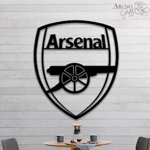 May include: Black metal Arsenal logo in a shield shape. The word "Arsenal" is above a cannon graphic. The logo is mounted on a white wooden background. The logo is a decorative item.