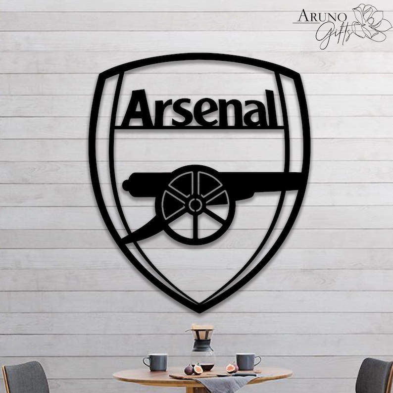 May include: Black metal Arsenal logo in a shield shape. The word "Arsenal" is above a cannon graphic. The logo is mounted on a white wooden background. The logo is a decorative item.