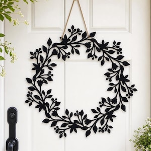 May include: A circular wall hanging featuring a black silhouette of interconnected leaves, stems, and small flowers. The piece is constructed from a flat material and is suspended by a thin, light brown twine loop attached to the top section.