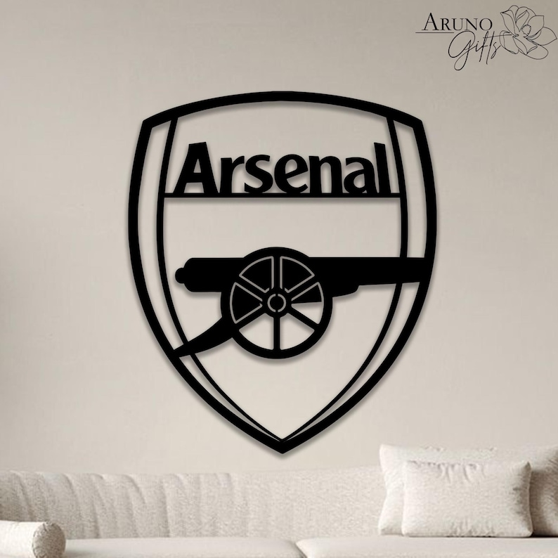 May include: Black metal Arsenal logo, featuring the team name above a cannon emblem, set against a white background. The logo is a shield shape, suitable for wall decor. The image is a close-up of the logo.