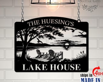 May include: Black metal lake house sign with the text "THE HUESING'S LAKE HOUSE". The sign features a lakeside scene with a tree, birds, and a dock with chairs. The sign is hanging from chains.