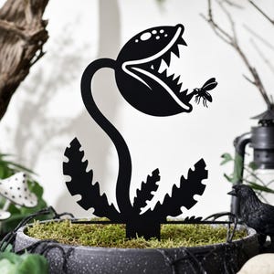 May include: A black metal silhouette of a carnivorous plant with a wide, open mouth featuring sharp teeth and a small fly positioned near the jaw. The plant has a curved stem and jagged leaves at the base, designed as a decorative garden stake.