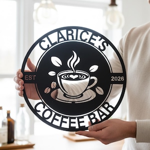 May include: Black metal coffee bar sign with the name "Clarice's" and the year "2026". The sign features a coffee cup design with steam and coffee beans. The sign is round and held by a person.