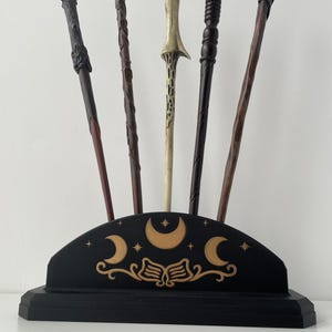May include: A collection of six decorative wands in various colors and designs, displayed in a black stand. The stand features crescent moon and star designs. The wands vary in length and have unique handles.