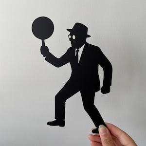 Può includere: Silhouette nera di un uomo in giacca e cravatta, cappello e occhiali che tiene in mano un oggetto rotondo. La figura cammina, e l'immagine è tenuta da una mano su sfondo bianco. La silhouette dell'uomo è un classico detective o spia.