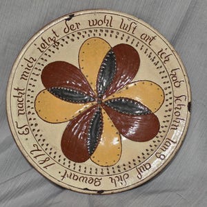 May include: A round, cream-colored ceramic plate with a floral design in brown, yellow, and gray. The plate has text around the edge in a dark brown script. The text includes the year 1812.