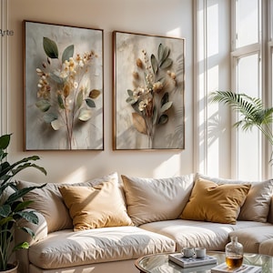 Könnte beinhalten: Zwei rechteckige gerahmte Wandbilder mit botanischen Illustrationen von getrockneten Blumen und Blättern in gedeckten Beige-, Braun- und Salbeigrüntönen. Die Kunstwerke hängen an einer hellen Wand über einem Sofa mit neutralen Kissen.