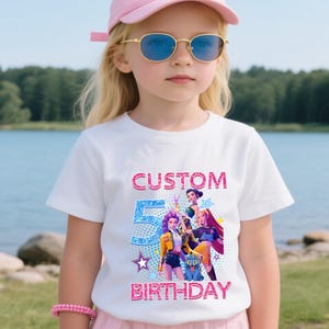 Puede incluir: Una camiseta blanca con un dise&ntilde;o gr&aacute;fico colorido para un 5&ordm; cumplea&ntilde;os. El dise&ntilde;o incluye las palabras "CUSTOM" y "BIRTHAY" en purpurina rosa, y el n&uacute;mero "5" en purpurina azul. El gr&aacute;fico presenta personajes de dibujos animados.