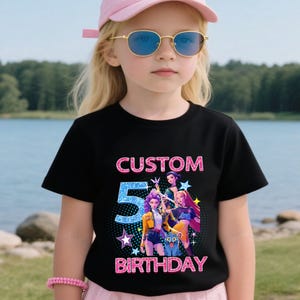 Puede incluir: Camiseta negra con un dise&ntilde;o gr&aacute;fico colorido con el texto "CUSTOM 5 BIRTHDAY". El gr&aacute;fico incluye personajes de dibujos animados y estrellas. La ni&ntilde;a de la imagen lleva una gorra rosa y gafas de sol.
