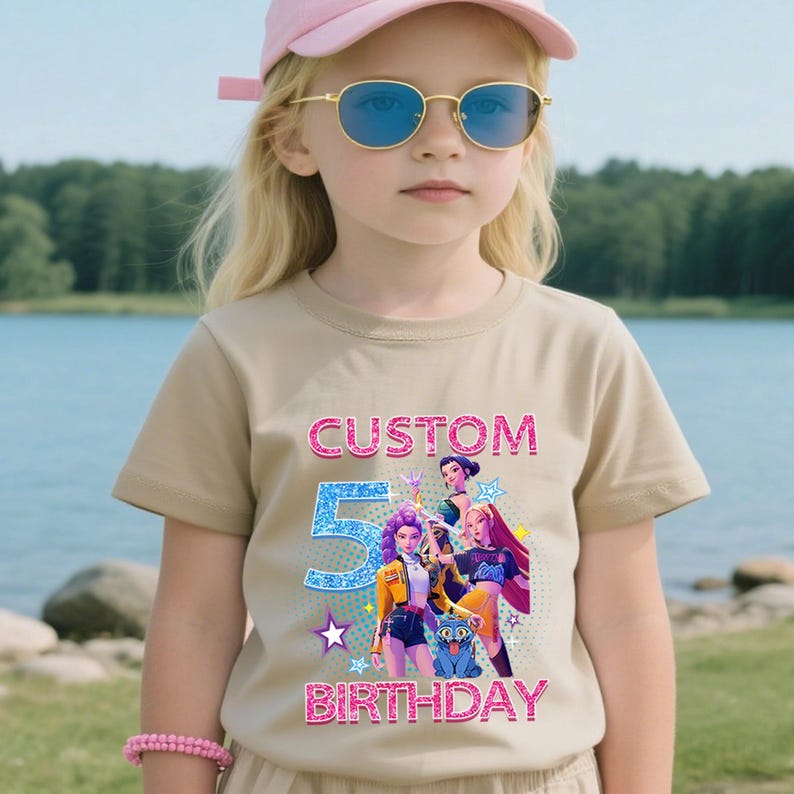 Puede incluir: Una camiseta color beige con las palabras "CUSTOM 5 BIRTHDAY" en purpurina rosa y azul. La camiseta presenta un gr&aacute;fico de personajes de dibujos animados. La persona de la imagen lleva una gorra rosa y gafas de sol.
