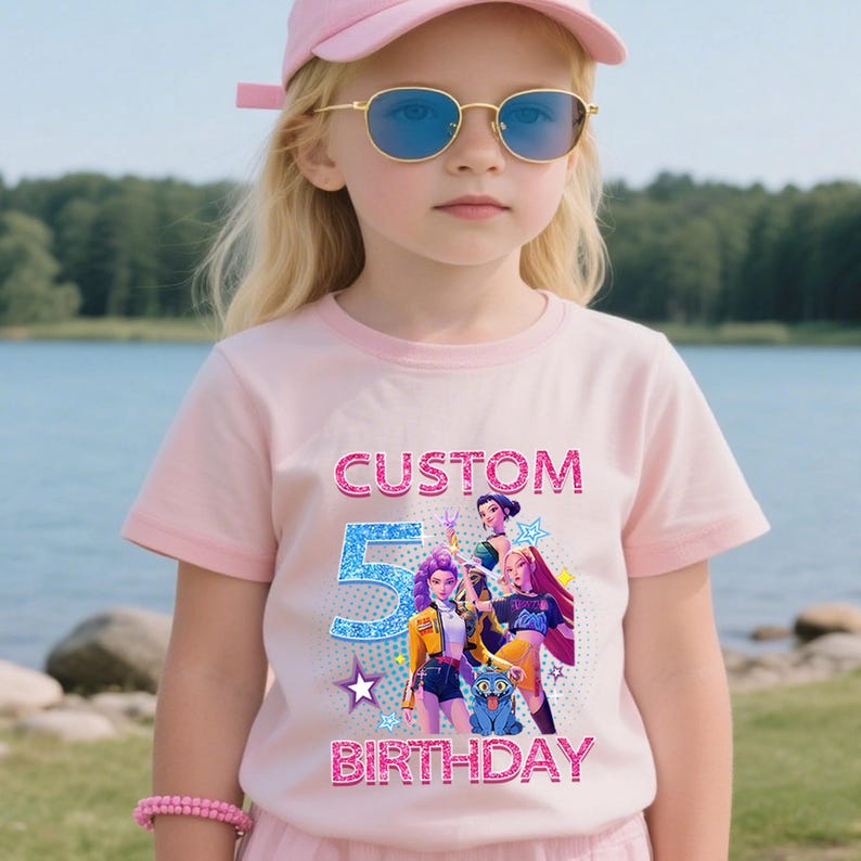 Puede incluir: Una camiseta rosa claro con un dise&ntilde;o gr&aacute;fico colorido. El dise&ntilde;o incluye el texto "CUSTOM 5 BIRTHDAY" y personajes de dibujos animados. La camiseta la lleva una persona joven con una gorra rosa y gafas de sol.