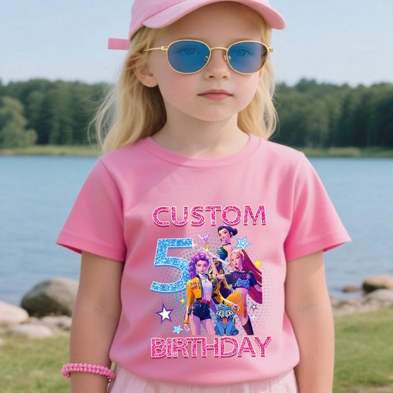 Puede incluir: Camiseta rosa de manga corta con el texto "CUSTOM 5 BIRTHDAY" en fuente brillante. La camiseta presenta un gr&aacute;fico colorido de personajes de dibujos animados. El ni&ntilde;o lleva una gorra y gafas de sol rosas.