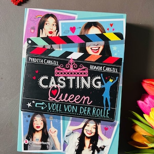 Könnte beinhalten: Ein Buchcover mit blauem Hintergrund und dem Titel "Casting Queen: Voll von der Rolle" in rosa und weißen Lettern. Das Cover zeigt Bilder von jungen Frauen mit verspielten Gesichtsausdrücken und einer Filmklappe.