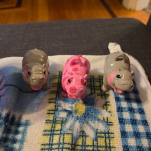 Może przedstawiać: Trzy małe figurki hipopotamów. Dwie są szare z różowymi akcentami, a jedna różowa z ciemniejszym różowym wzorem. Ułożone są na niebiesko-żółtej wzorzystej tkaninie.