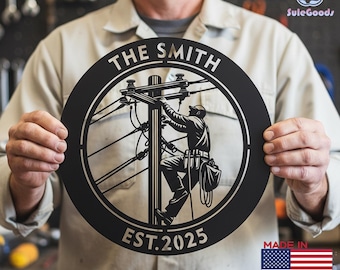 May include: A black metal circular sign featuring a lineman working on a power pole. The sign is personalized with "THE SMITH" and "EST.2025" in white text. The sign is held by a person. The sign is made in the USA.