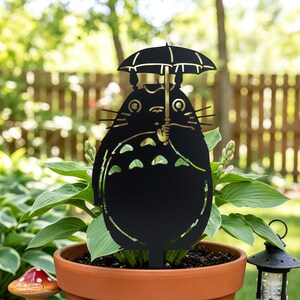 May include: Black metal garden stake featuring a cartoon character holding an umbrella. The character has large eyes and a round body. The stake is placed in a terracotta pot with green plants, with a blurred background of a wooden fence and greenery.