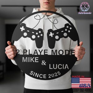 May include: Black metal wall art featuring two game controllers connected by hearts. The text reads "2 PLAYE MODE MIKE & LUCIA SINCE 2025". The art is a circular design, perfect for a game room or as a personalized gift.