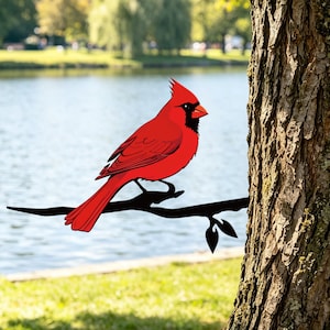 May include: A bright red cardinal bird with a black face mask and crest, perched on a black metal branch silhouette. The bird features detailed wing feathers and a pointed beak, designed as a decorative garden ornament attached to a tree trunk.